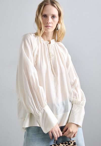 Blusa di seta crema con scollatura arricciata, maniche lunghe e voluminose, polsini con bottoni e una texture leggermente trasparente. Indossata con jeans e una pochette con motivo.