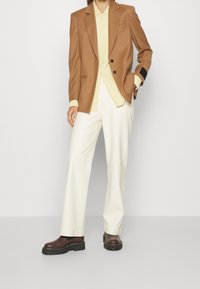 Brauner maßgeschneiderter Blazer, gelbes Hemd mit Knöpfen, weiße weite Hose und braune klobige Stiefel mit schwarzer Sohle. Glattes Material und strukturiertes Design.