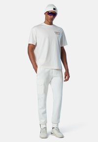 Hombre con gorro blanco, gafas de sol moradas, camiseta blanca de North Sails, pantalones de chándal blancos con bolsillo y zapatillas blancas sobre fondo plano.