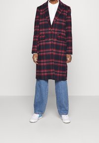 Henkilö, jolla on päällään pitkä punamusta ruudullinen takki valkoisen paidan, vaaleansiniset farkut ja valkoiset tennarit sinisillä yksityiskohdilla, seisoo harmaalla lattialla.