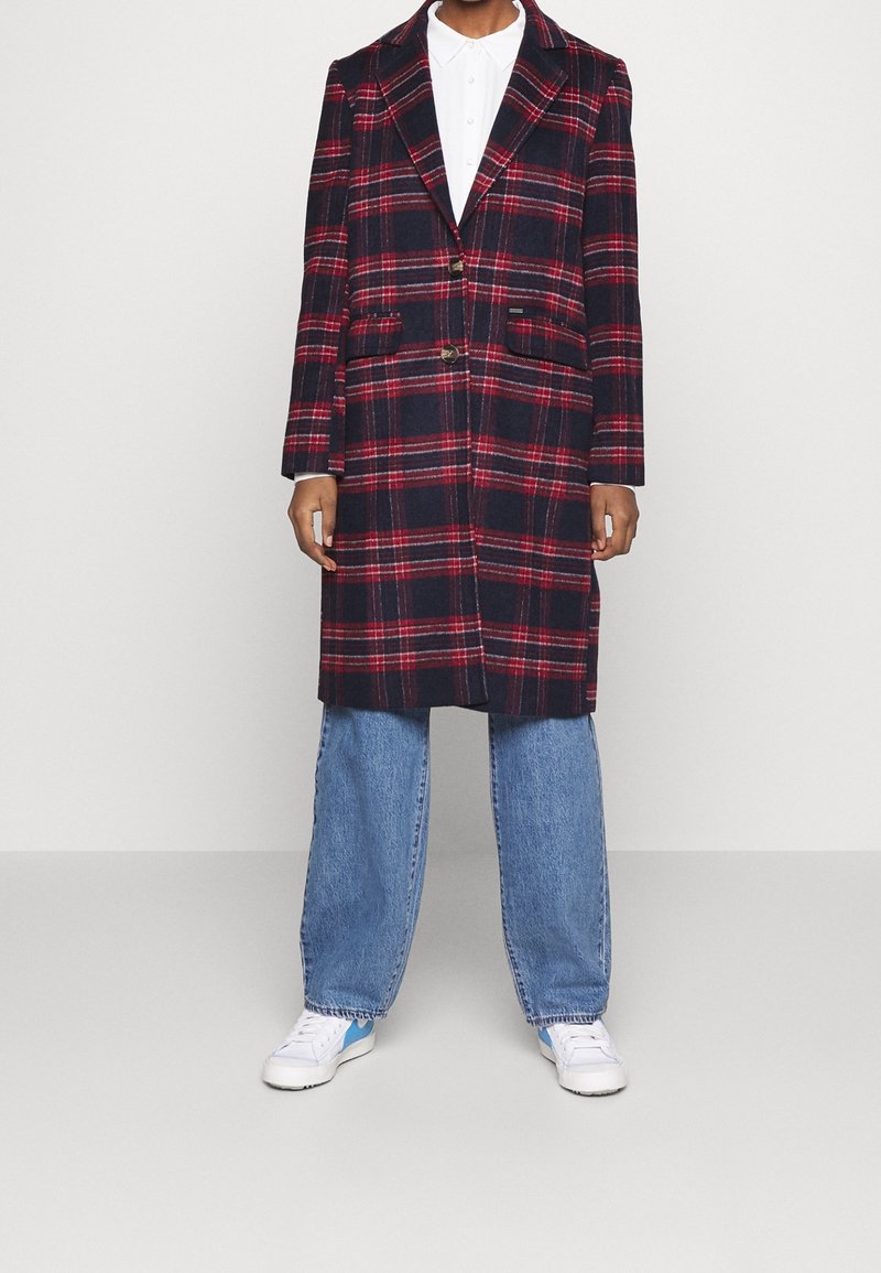 Henkilö, jolla on päällään pitkä punamusta ruudullinen takki valkoisen paidan, vaaleansiniset farkut ja valkoiset tennarit sinisillä yksityiskohdilla, seisoo harmaalla lattialla.