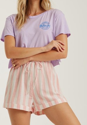 Kvinde iført en kort lavendel t-shirt med blåt logo og lyserøde og hvide vertikale stribede shorts med snor i taljen, armene krydsede.