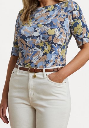 Kvinde iført en blå, beige og grøn blomsterprintet kortærmet top, stoppet ned i hvide bukser med høj talje og et brunt bælte. Den ene hånd i lommen.