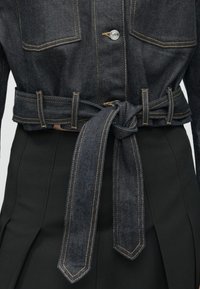 Chaqueta de mezclilla oscura con cintura ceñida, costuras amarillas distintivas, dos bolsillos delanteros y cierre de botones a presión. Combinada con una falda negra.