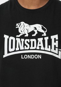 Must värviline t-särk on valmistatud puuvillast, millel on suur valge lõvijoonistus ja "LONSDALE LONDON" tekst paksus kirjas rinna kohal.
