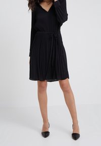 Robe plissée noire avec un décolleté en V, des manches longues et une taille à nouer. Tissu lisse avec un design fluide, associée à des talons aiguilles noirs.