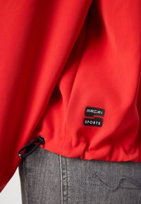 Rode sportjacket met elastische zoom en verstelbaar koord. Bevat een zwart label met het "Marc Cain SPORTS" logo. Gladde, lichte stof.