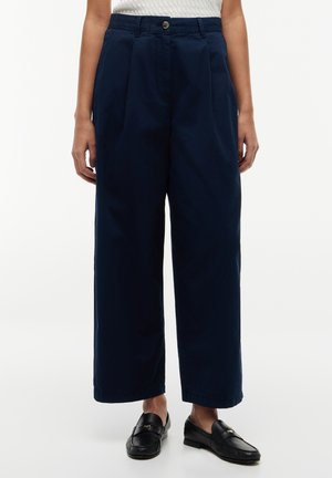 Personne portant un pantalon large bleu marine, un haut sans manches texturé blanc et des mocassins en cuir noir, debout sur un fond blanc.