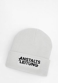 White knitted beanie with a ribbed texture featuring a rectangular patch that displays the text "ANSTALTSLEITUNG" in bold black letters.
