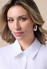 Biała koszula z kołnierzykiem ma subtelny połysk i dekoracyjny guzik. Modelka nosi srebrne kolczyki w kształcie kół, ma falujące włosy i makijaż w neutralnych odcieniach.