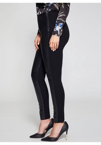 Černé legíny s přiléhavým střihem, zdobené lesklým panelem podél vnějšího švu, v kombinaci s černými špičatými podpatky.