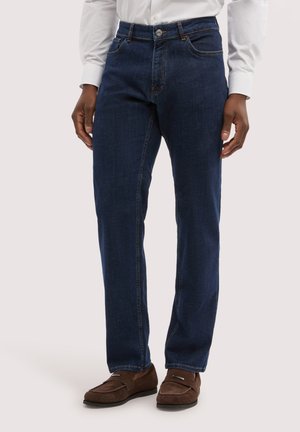 Mann trägt dunkelblaue Jeans mit geradem Bein, weißes Hemd mit Knöpfen und braune Slip-On-Loafer, steht vor einem schlichten Hintergrund.