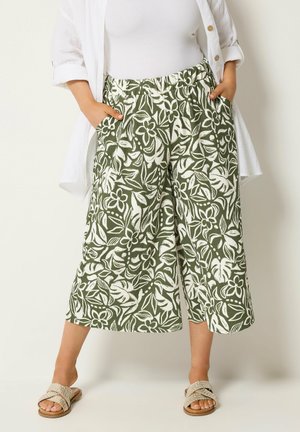 Kobieta w zielonych szerokich spodniach 7/8 z białym tropikalnym nadrukiem liści i kwiatów, białym topie, otwartej białej koszuli i beżowych sandałach.