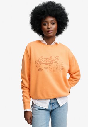 Frau mit lockigem Haar, die einen orangen Superdry-Sweatshirt über einem weißen Hemd mit Knöpfen und hellblauen Jeans trägt, steht vor einem einfarbigen Hintergrund.
