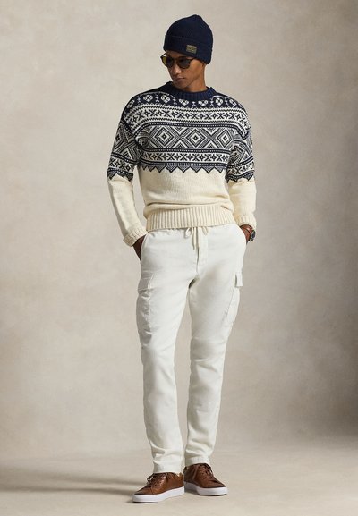 Πλεκτό πουλόβερ με ναυτικό μπλε, κρεμ και γεωμετρικά σχέδια· κρεμ παντελόνι cargo· καφέ αθλητικά παπούτσια· ναυτικό μπλε σκούφος· στρογγυλά γυαλιά ηλίου.