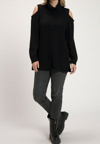 Svart oversize tröja med flätmönster, hög hals och öppningar för kalla axlar, i kombination med mörka jeans och kraftiga svarta stövlar.