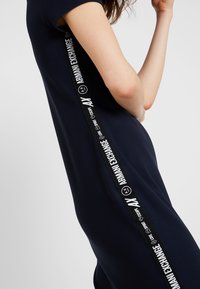 Granatowa sukienka z krótkimi rękawami, z czarnym, markowym paskiem na boku z białym tekstem i szczegółami logo. Gładka, dopasowana tekstura.