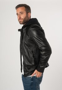 Zwarte leren jack met een gestructureerde afwerking, ritszakken en een gebreide hoodie eronder. Heeft een getailleerd ontwerp en zichtbare stikdetails.