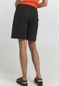 Schwarze Shorts mit glatter Textur, ausgestattet mit einem Taillenbund in mittlerer Höhe und einer Gesäßtasche. Kombiniert mit schwarzen Sandalen ist das Outfit minimalistisch und funktional.
