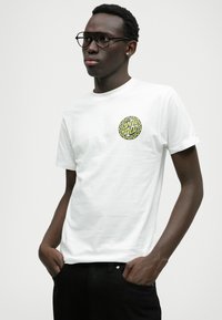 Balts īsām piedurknēm T-krekls ar apaļu grafisko logotipu dzeltenā un melnā krāsā uz krūts, apvienots ar melnām biksēm.