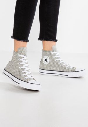 Osoba nosząca jasnoszare siateczkowe trampki Converse na wysokiej cholewce z białymi sznurowadłami i białą podeszwą z czarną lamówką, w połączeniu z czarnymi spodniami z postrzępionym dołem.