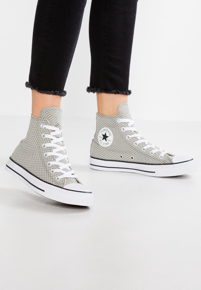 Personne portant des baskets montantes Converse en mesh gris clair avec des lacets blancs et des semelles blanches bordées de noir, assorties à un pantalon noir à ourlet effiloché.