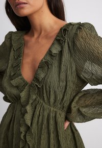 Robe vert olive avec un décolleté en V profond, un col à volants et un tissu texturé et froissé. Manches longues avec un subtil accent de volants aux poignets.