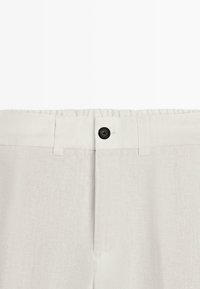 Ceinture d'un pantalon en lin beige avec un bouton noir et une boutonnière, montrant des passants pour ceinture et une fermeture éclair dissimulée.