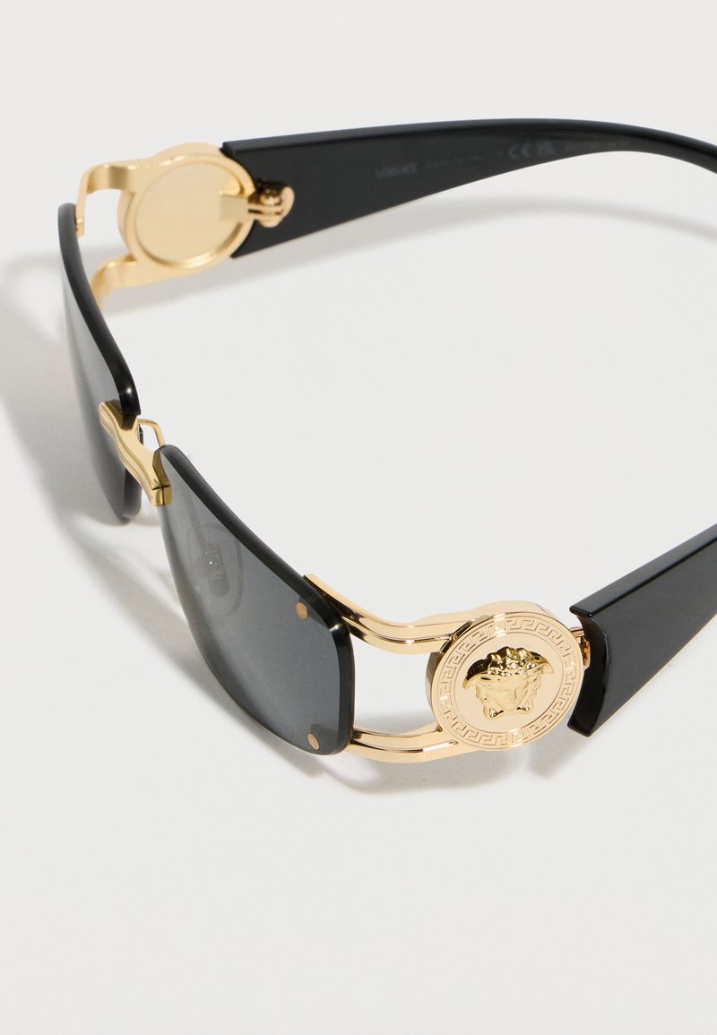 Lunettes de soleil rectangulaires noires avec accents dorés et emblème de Méduse sur les branches, montrant les détails de côté et de face.