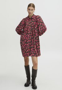 Abito camicia nero a fiori con motivi di fiori rosa e verdi, maniche lunghe, colletto con bottoni e una vestibilità comoda. Abbinato a stivali neri al ginocchio.