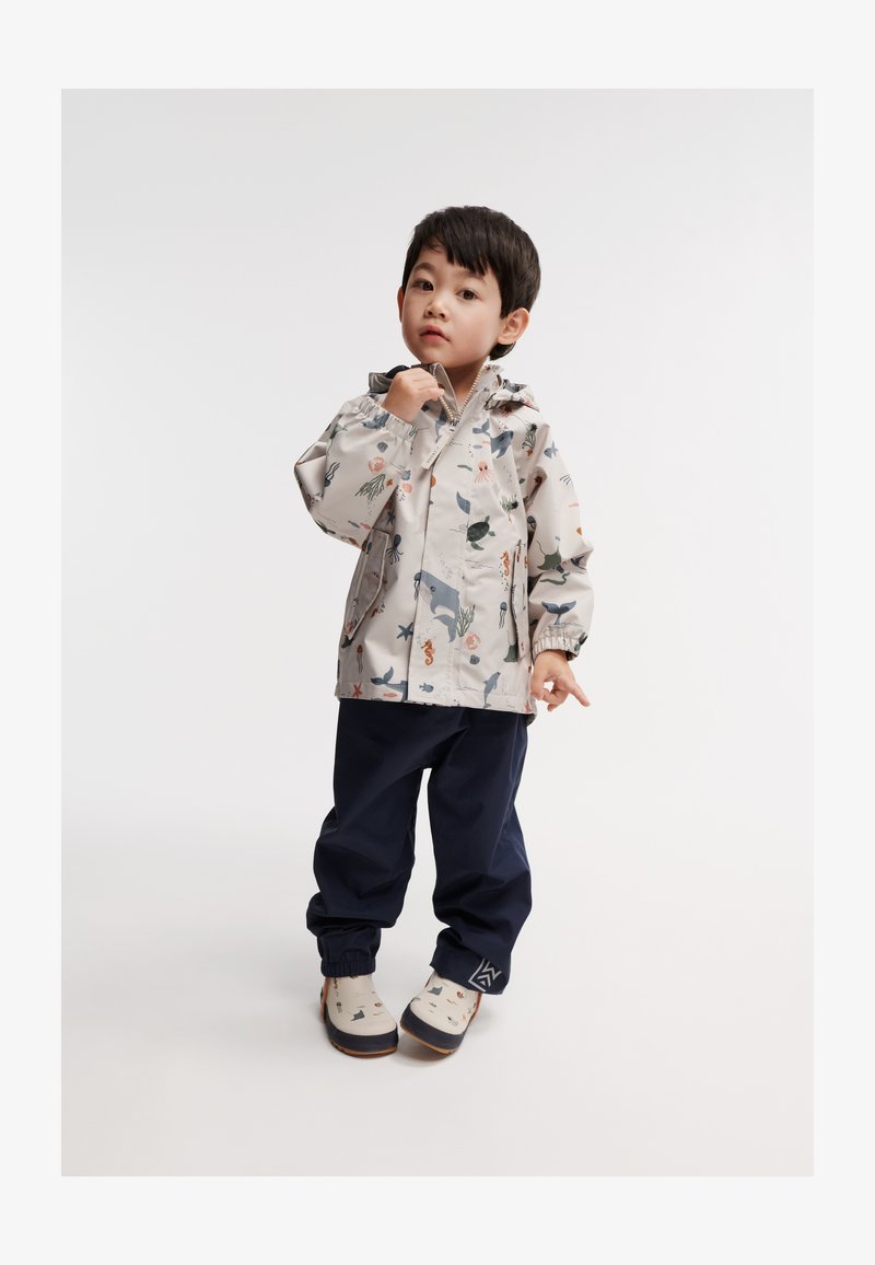 Veste imperméable beige clair avec des motifs marins, associée à un pantalon bleu marine et des chaussures à motifs. Elle possède une capuche et des poignets élastiques.