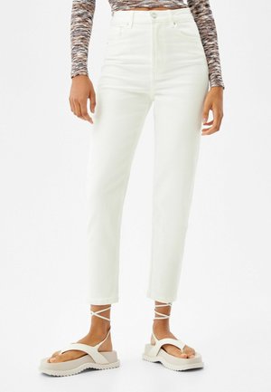 Persoon gekleed in een wit recht model met hoge taille jeans, een meerkleurige longsleeve en witte plateauzandalen met enkelbandjes.