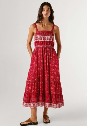 Mujer de pie con un vestido midi sin mangas rojo con estampados florales y sandalias marrones, con las manos en los bolsillos, fondo neutro.