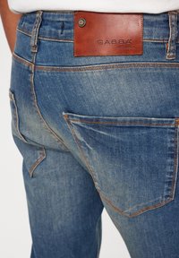 Ljusblå denimjeans med en lädermärkeslapp, synliga sömdetaljer och flera fickor, inklusive en distinkt design på bakfickan.
