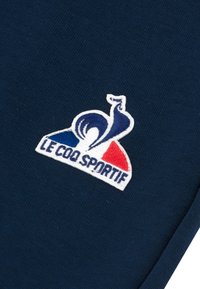 Tessuto blu navy con un logo ricamato bianco raffigurante un gallo, dettagli rossi e blu, e il testo "LE COQ SPORTIF" sotto il logo.