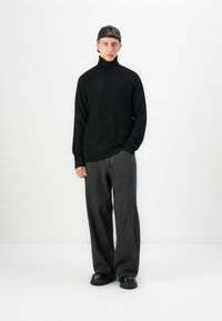 Jonge man in een zwart coltrui, loszittende donkergrijze jeans, zwarte chunkyschoenen en een achterstevoren gedragen zwarte pet op een witte achtergrond.