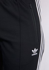 Pantaloni sportivi neri in tessuto testurizzato, dotati di una tasca con zip e tre strisce bianche lungo il lato, con logo Adidas bianco.