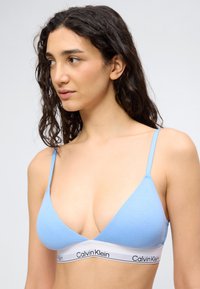 Γυναίκα με μακριά σγουρά σκούρα μαλλιά, φορώντας ένα ανοιχτό μπλε bralette Calvin Klein με λεπτές τιράντες, μπροστά σε ανοιχτό γκρι φόντο.