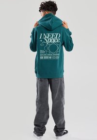 Vihreä huppari, jossa on grafiikkaprintti selässä ja teksti "Tarvitsen tilaa" valkoisella, yhdistettynä harmaisiin löysiin housuihin ja tennareihin.