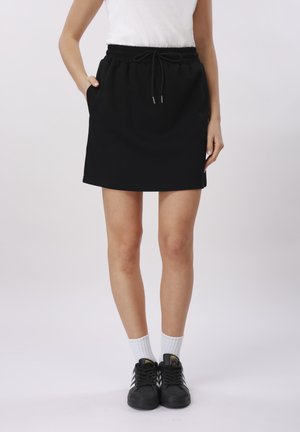 Femme portant une jupe noire à cordon, des chaussettes blanches, des baskets noires Adidas et un haut blanc sans manches, debout avec les mains partiellement dans les poches.