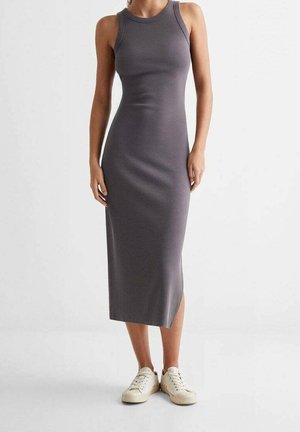 Femme portant une robe midi grise ajustée sans manches, associée à des baskets blanches, debout devant un fond clair uni.