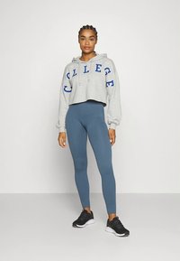 Felpa grigia con cappuccio tagliata, con scritta blu "COLLEGE" e cordini grigi, abbinata a leggings blu e scarpe sportive nere.