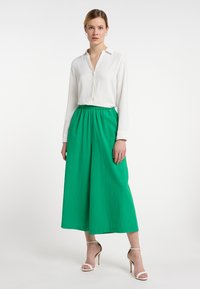 Witte blouse met een kraag, gecombineerd met groene broeken met hoge taille en brede pijpen. De schoenen zijn beige met een dunne band. Gladde stoffen, minimalistisch ontwerp.