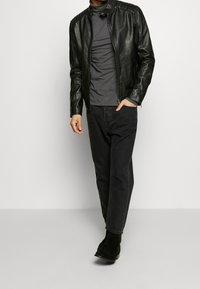 Man wearing black leather jacket, dark gray turtleneck, black jeans, and black suede boots, standing against a plain light background.