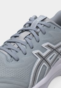 Ανοιχτό γκρι παπούτσι τρεξίματος Asics με υφασμάτινη υφή, παχιά επίπεδα κορδόνια και λογότυπο της μάρκας στο πλάι και στην γλώσσα.