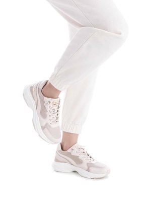 Zapatillas beige con parte superior texturizada, paneles de malla y suela acolchada. Presentan cordones, detalle del logo y costuras de acento. Combinadas con pantalones de chándal blancos.