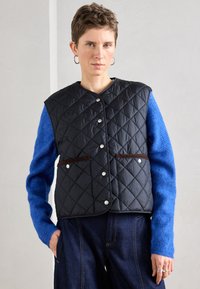 Piros fekete steppelt mellény patentgombokkal, barna díszítésekkel és két elülső zsebbel, élénk kék kötött pulóverre rétegezve.