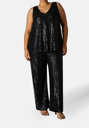 Person mit Übergröße trägt ein schwarzes ärmelloses Paillettenoberteil und passende, weit geschnittene Paillettenhose sowie goldene offene Absätze.