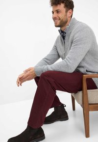 Maglione grigio con zip e polsini a costine, pantaloni bordeaux e stivaletti neri, seduto su una sedia, con una finitura in tessuto testurizzato.