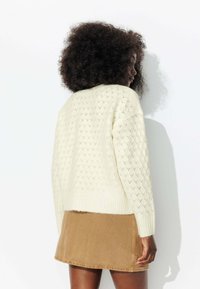 Pull en tricot crème avec un motif en losanges texturés, col rond et poignets côtelés, associé à une jupe en daim marron.